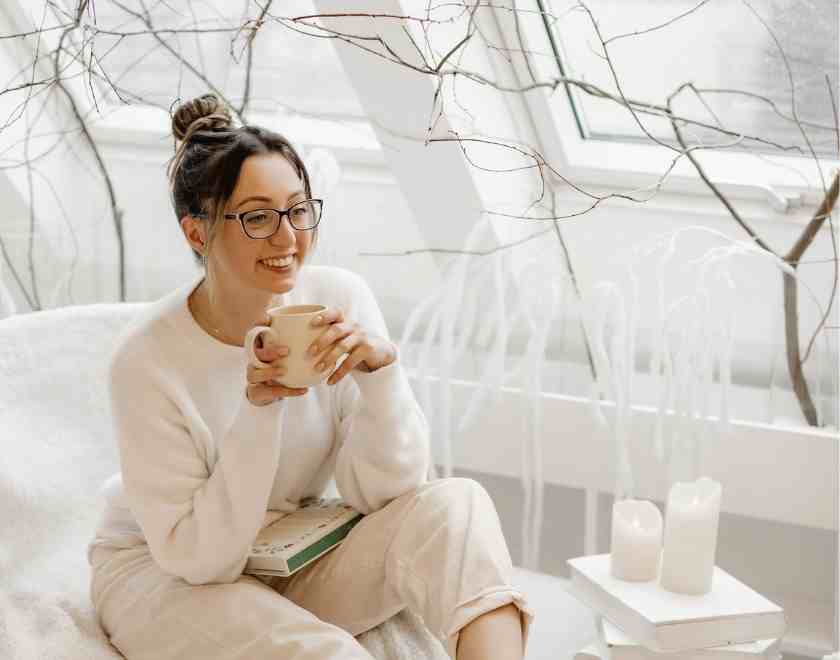 Woman with glasses and messy bun sitting on bed with book and candles focusing on her word of the year
